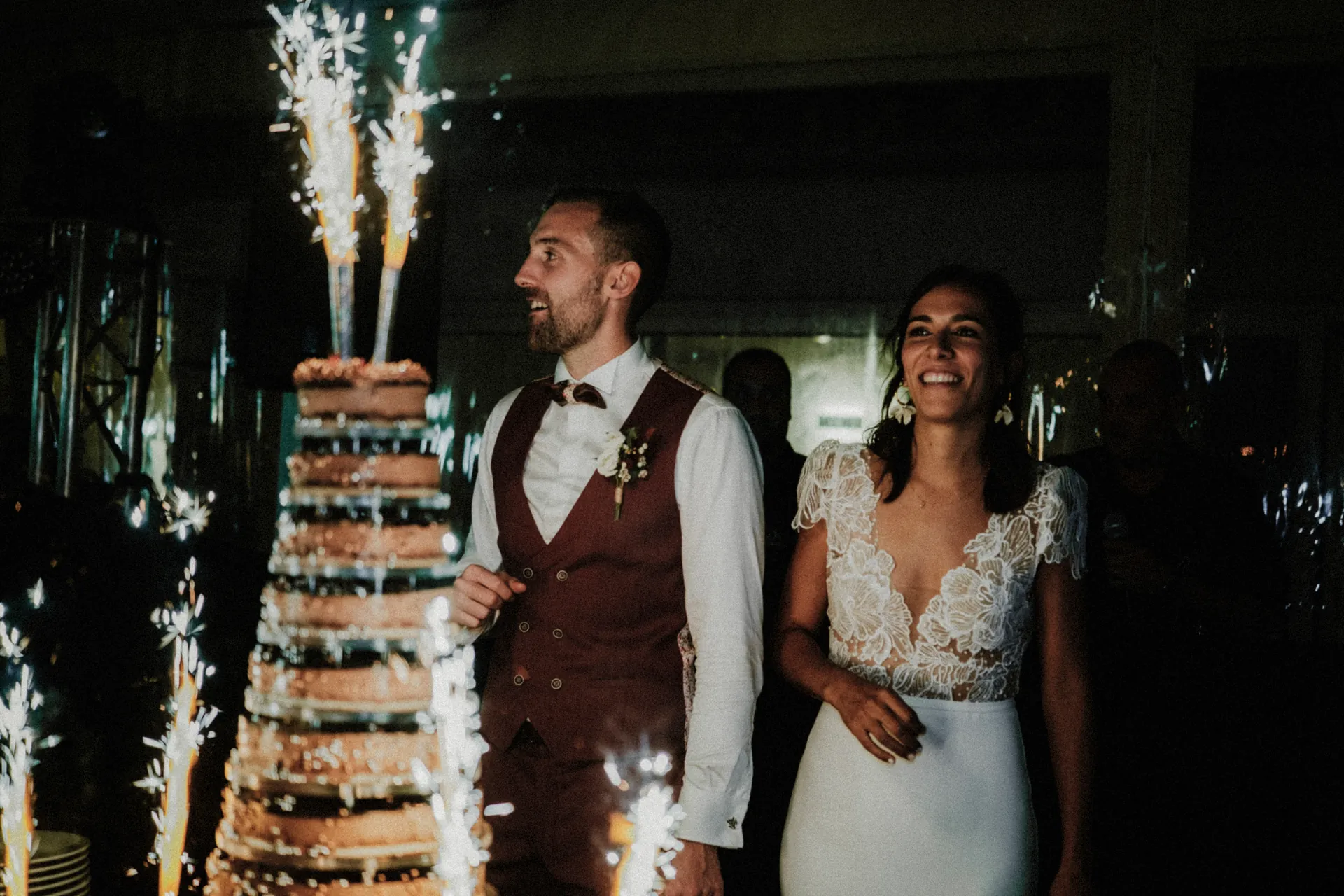 Un couple se tient à côté d'un grand gâteau à plusieurs étages orné de cierges magiques, capturé dans les moindres détails par le photographe de mariage Jeremy Dan, basé à Sceaux. L'homme porte un gilet et un nœud papillon, tandis que la femme est éblouissante dans une robe en dentelle.