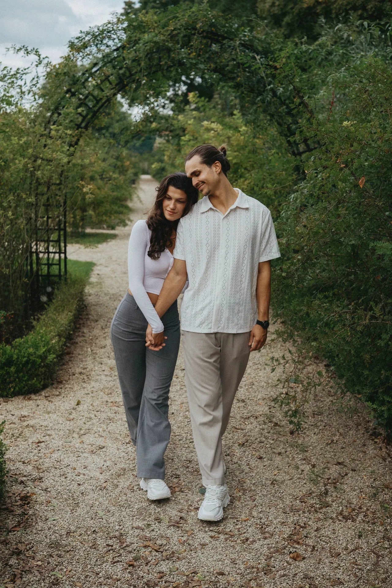 Un couple marche main dans la main sur un chemin de gravier entouré de verdure, avec une arche au-dessus de la tête, capturé à travers l’objectif de Jeremy Dan, photographe de mariage basé à Sceaux, Hauts-de-Seine.