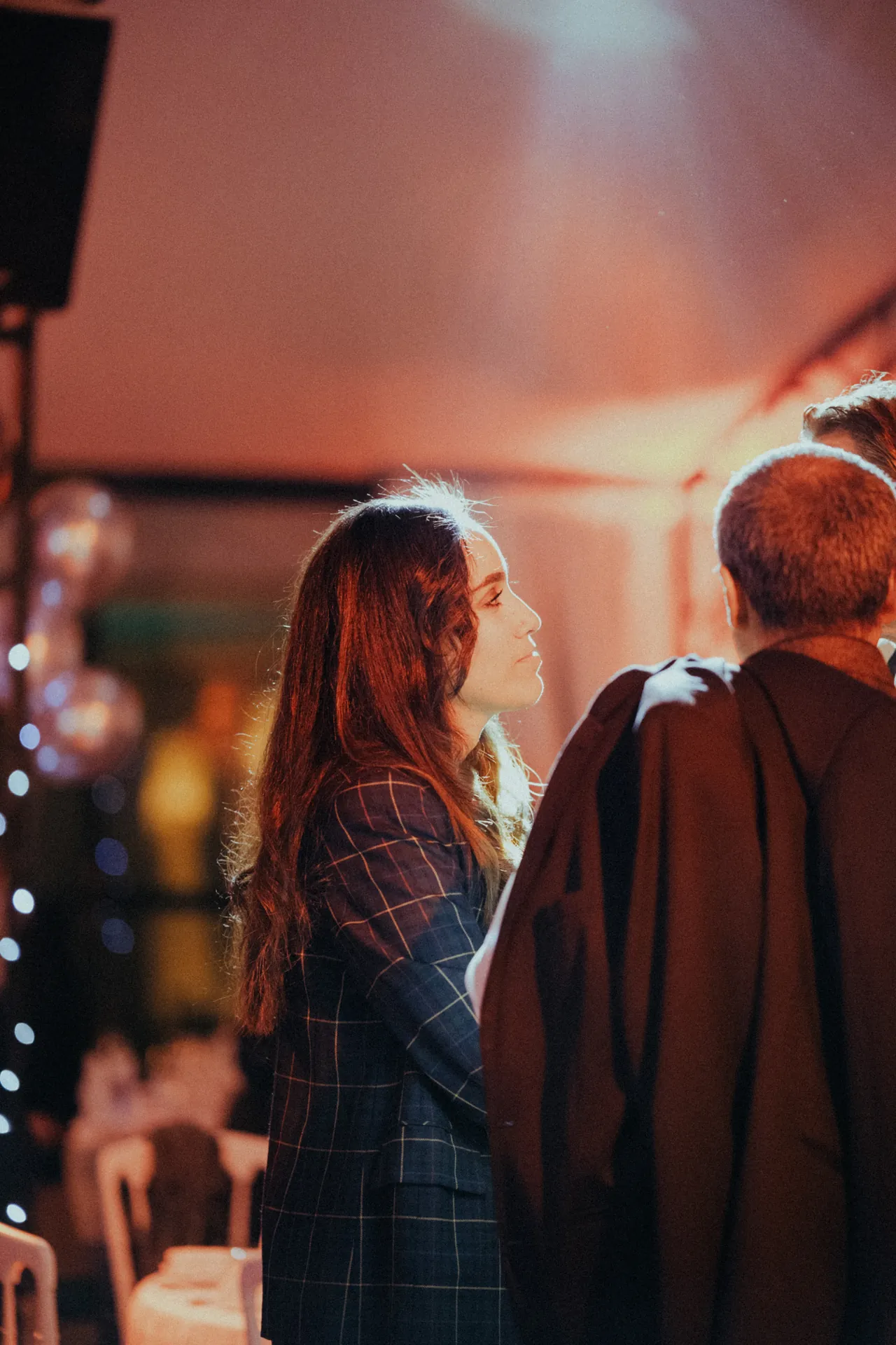Une femme en blazer à carreaux discute avec un de ses collègues durant une soirée d'entreprise. Ballons et guirlandes lumineuses ajoutent une touche de fantaisie à l'arrière-plan. Capturé par Jeremy Dan, photographe et vidéaste d'entreprise à Sceaux, Hauts-de-Seine.