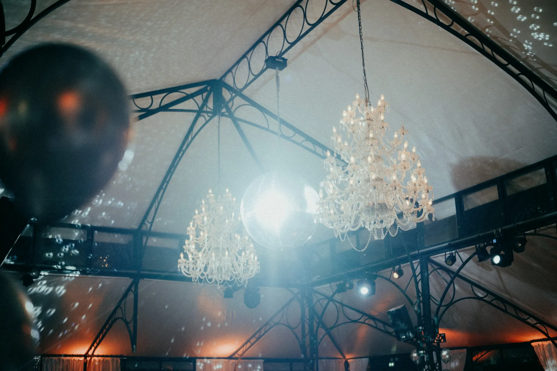 Des lustres ornés et une boule disco sont suspendus au plafond à l'intérieur d'une tente, avec un éclairage décoratif créant une atmosphère festive. Capturé par Jeremy Dan, photographe et vidéaste d'entreprise basé à Sceaux, dans les Hauts-de-Seine.