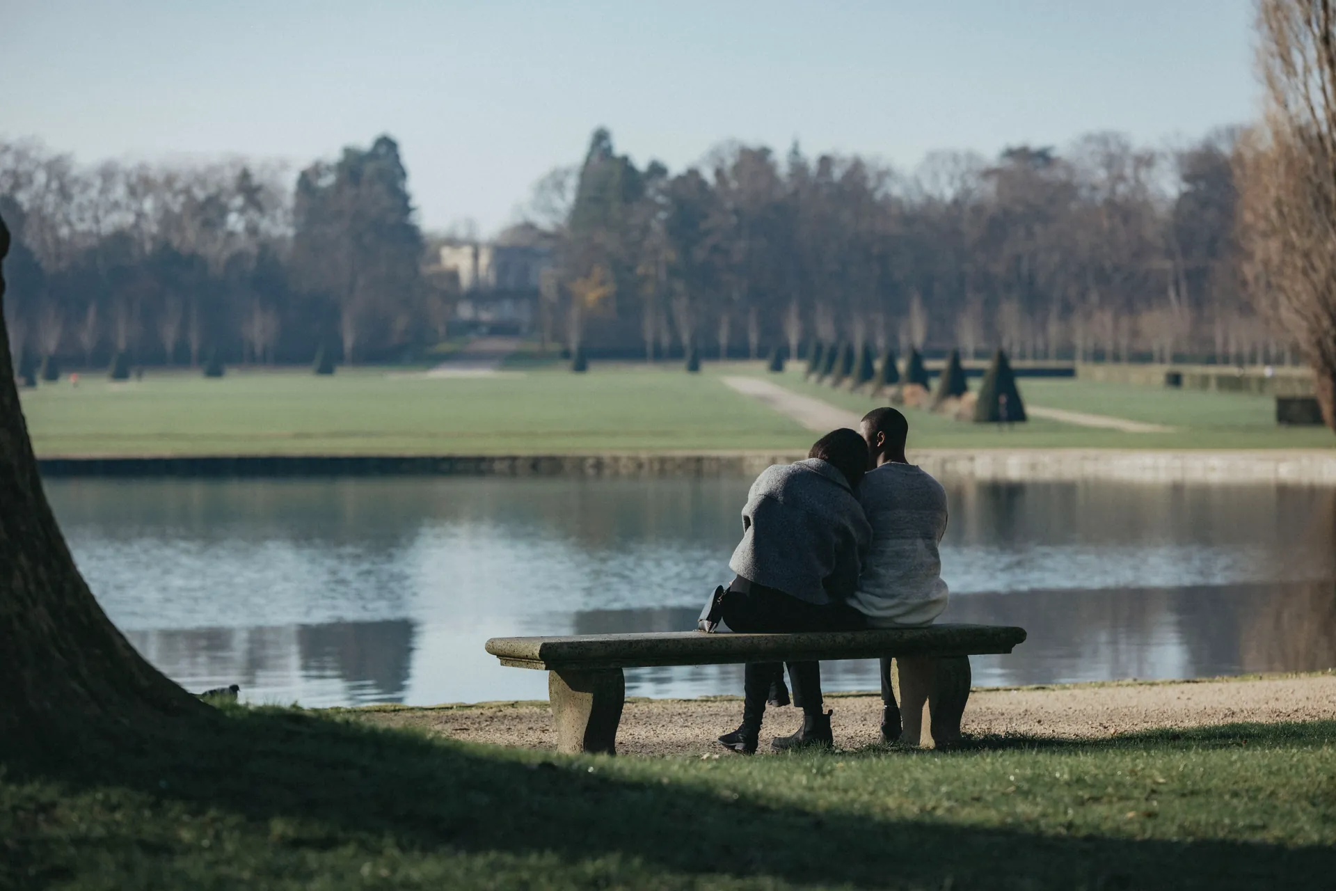 Un couple est assis face à un lac dans un parc. La femme pose sa tête sur l'épaule de l'homme et ils regardent l'horizon. Photo réalisée dans le cadre d'une séance photos de couple couverte par Jeremy Dan, photographe de couple basé à Sceaux.