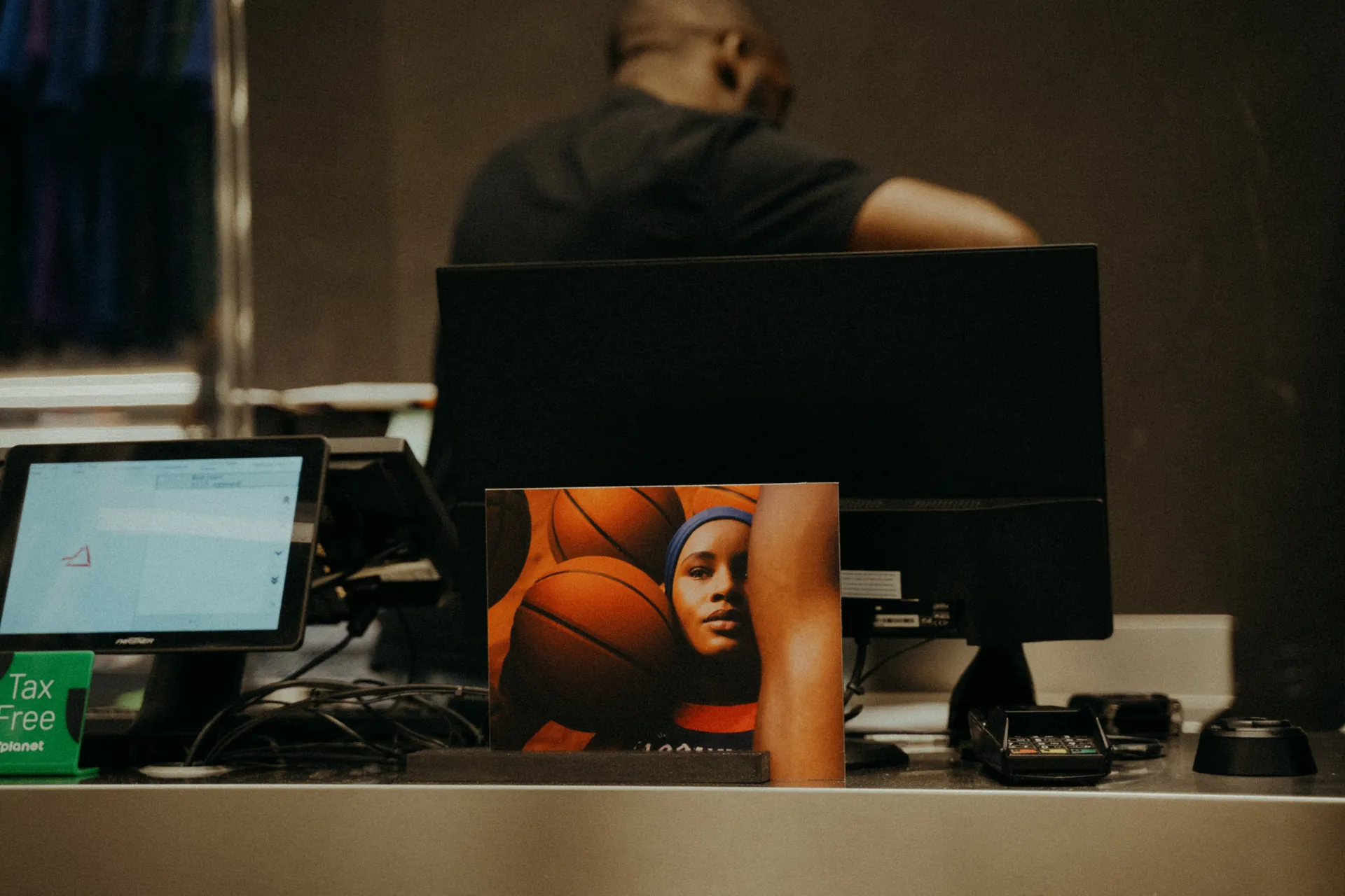 Un organisateur d'événement se tient à une caisse en arrière plan. Au premier plan, une photo montre un enfant avec des ballons de basket à côté de l'équipement. Capturé par Jeremy Dan, photographe et vidéaste événementiel à Sceaux, Hauts-de-Seine.