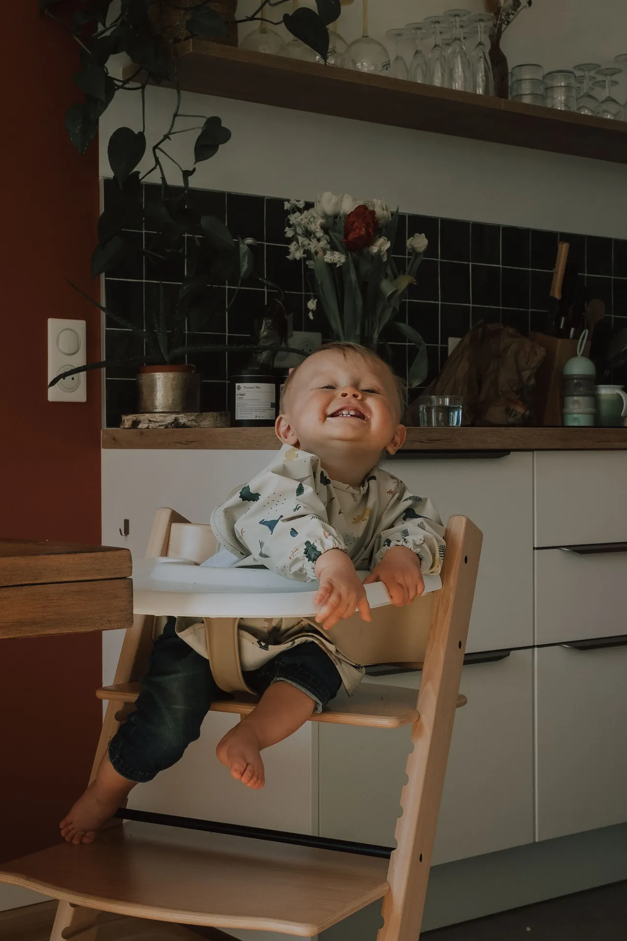 Dans une cuisine, un bébé est assis joyeusement dans une chaise haute, les yeux clos de bonheur. Derrière lui, des étagères regorgent d'ustensiles de cuisine variés. Capturé par Jeremy Dan, photographe de famille à Sceaux, Hauts-de-Seine.