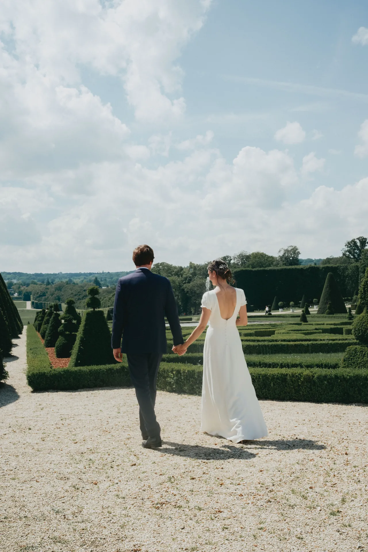 Le marié et la mariée marchent main dans la main au parc de Sceaux en tenue de mariage durant leur séance photos de couple.