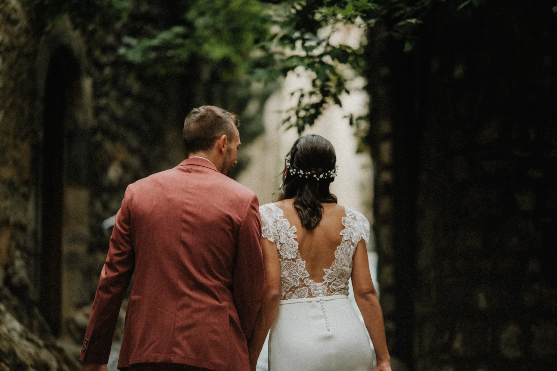Un couple de mariés se promène en tenue de mariage dans un petit village près de Montpellier, la femme en robe blanche et l'homme en costume rouge, entourés d'arbres et de murs en pierre. Capturé par Jeremy Dan, photographe et vidéaste de mariage basé à Sceaux, Hauts-de-Seine.