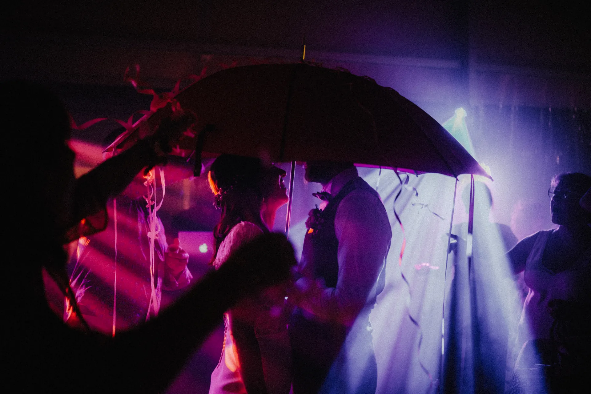 Le marié et la mariée se tiennent sous un parapluie, entourés de lumières colorées et de confettis, créant une atmosphère festive lors de la première danse de leur mariage. Capturé par Jeremy Dan, photographe de mariage basé à Sceaux, dans les Hauts-de-Seine.