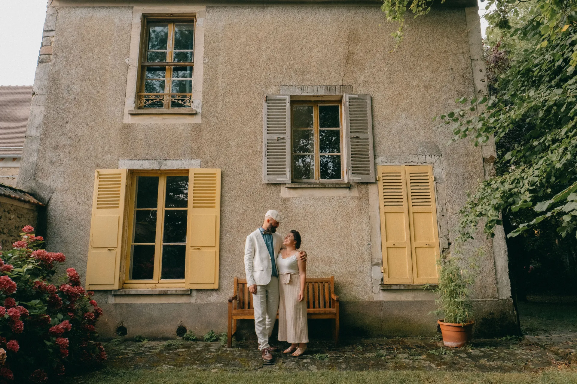 Un couple se tient côte à côte devant une vieille maison en pierre aux volets jaunes, entourée de verdure et de plantes en pot. Capturé par Jeremy Dan, photographe de mariage basé à Sceaux.