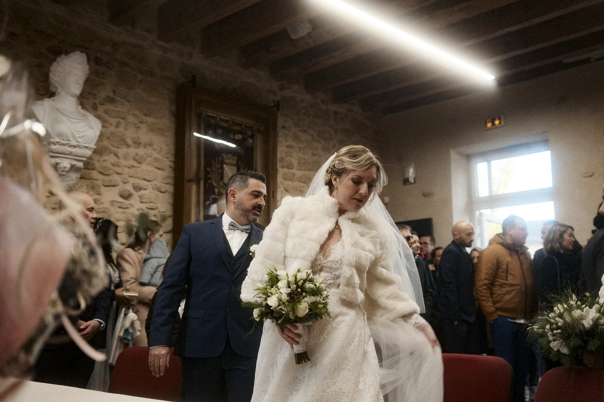 Une mariée et un marié en tenue de mariage marchent à l'intérieur de la mairie parmi des invités assis. La mariée tient un bouquet et porte une étole de fourrure blanche et un voile, tandis que le marié porte un costume. Capturé par Jeremy Dan, photographe et vidéaste de mariage basé à Sceaux, dans les Hauts-de-Seine.
