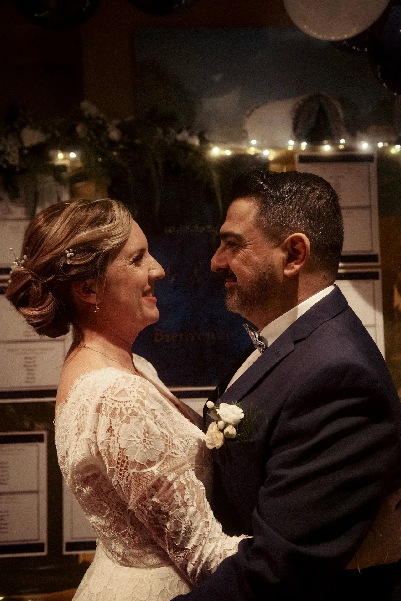 Un couple de mariés en tenue de soirée se fait face, souriant, dans un cadre intérieur magnifiquement éclairé et fleuri. Capturé par Jeremy Dan, photographe et vidéaste de mariage basé à Sceaux, Hauts-de-Seine.
