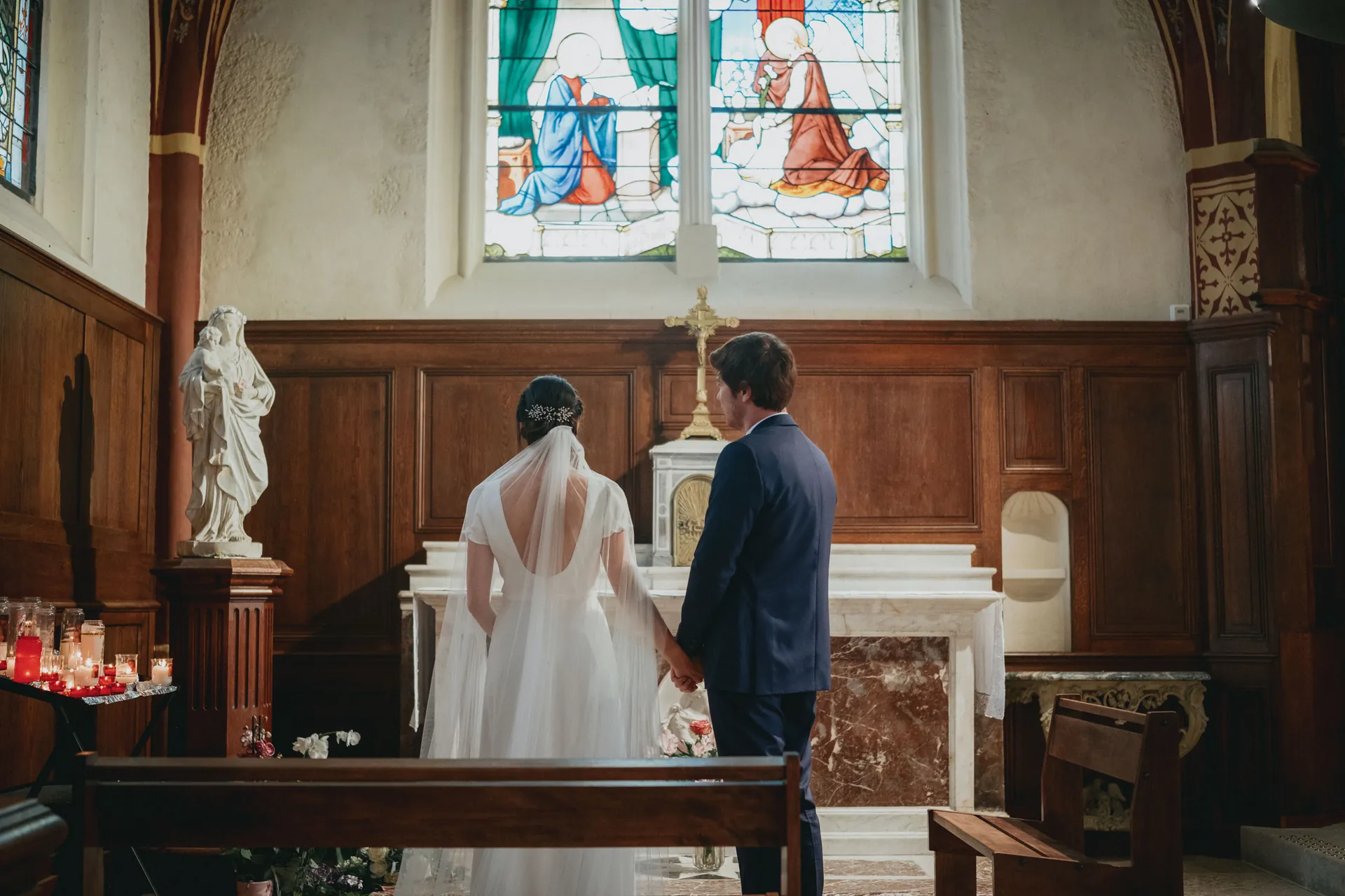 Un couple en tenue de mariage se tient la main dans une chapelle, face à un autel sous des vitraux. Capturé par Jeremy Dan, photographe et vidéaste de mariage.