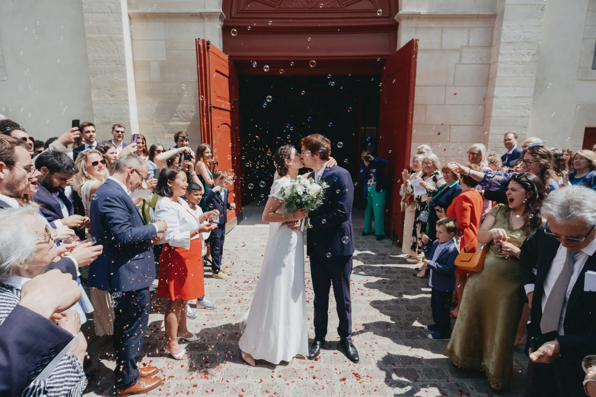 Un couple de mariés s'embrasse devant l'église de Sceaux, entourés d'invités joyeux et de bulles flottant dans l'air. Capturé par Jeremy Dan, photographe de famille à Sceaux, Hauts-de-Seine.
