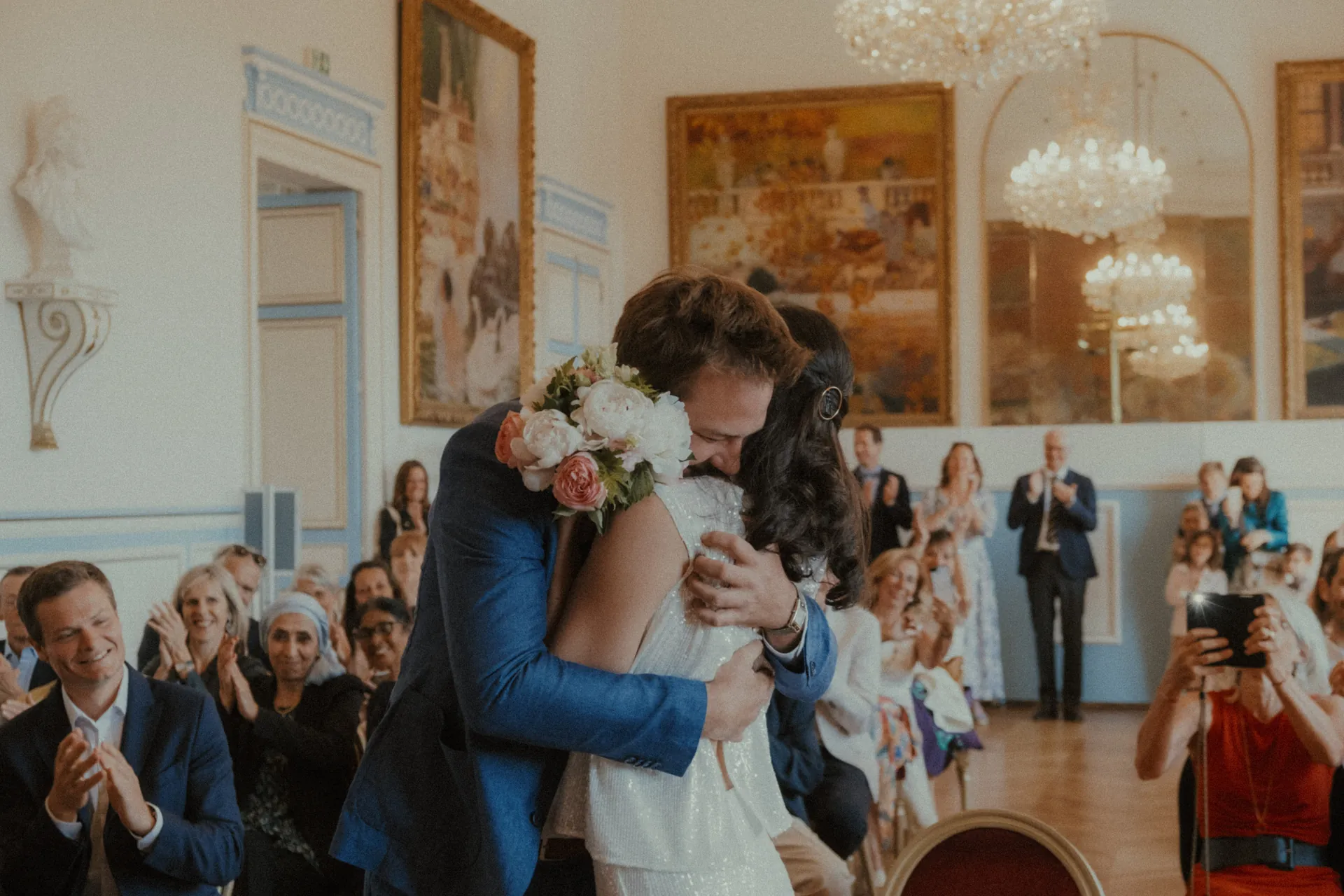 Les mariés s'embrassent dans une salle de l'hôtel de ville décorée et remplie d'invités assis. Alors que les gens applaudissent et que certains prennent des photos, de grands tableaux et des lustres plantent le décor. Capturé magnifiquement par Jeremy Dan, photographe de mariage basé à Sceaux, ce moment résonne des échos de l'amour.