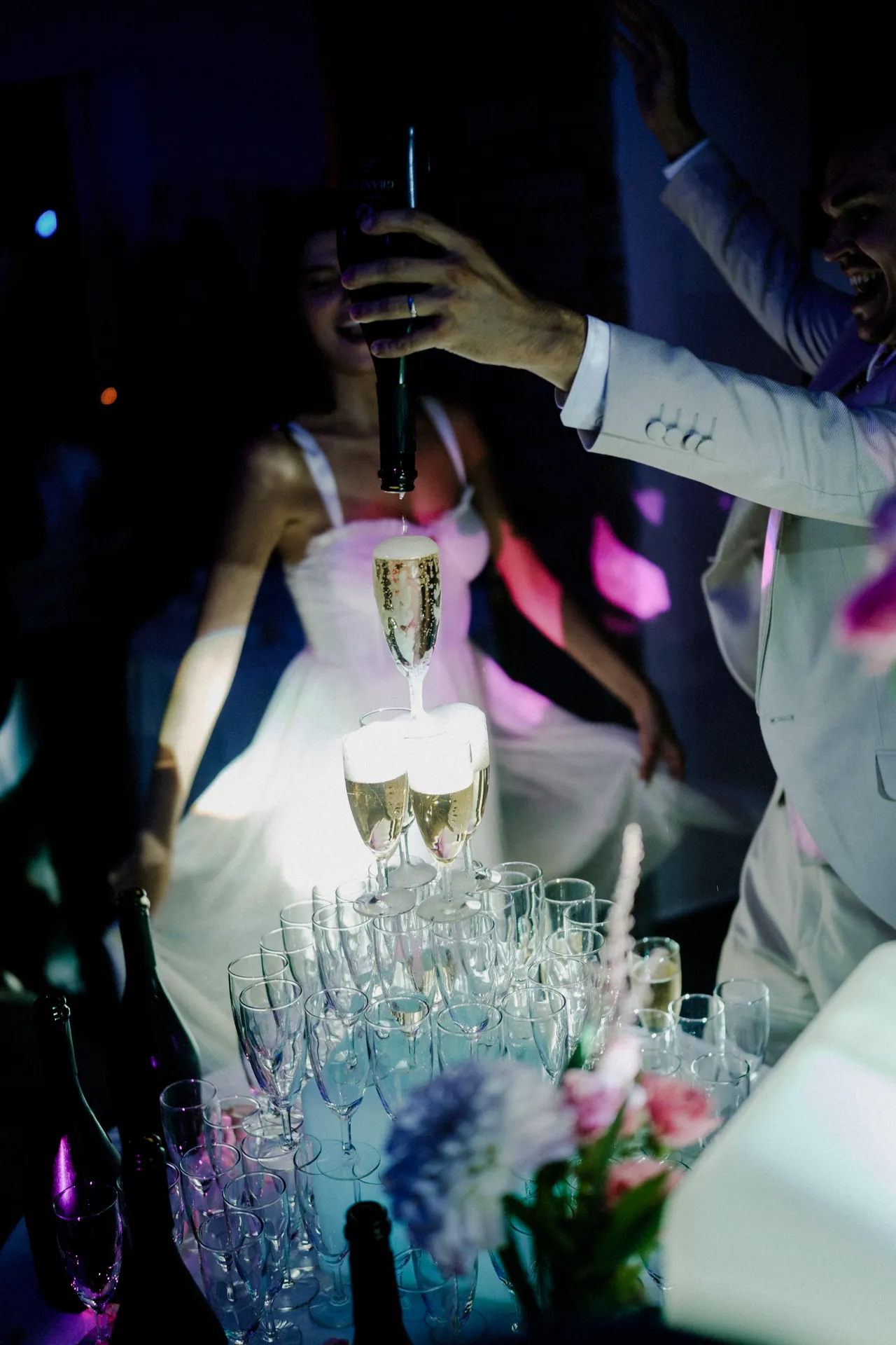 La marié verse du champagne dans une tour de verre lors de la soirée de son mariage, entourée de fêtards et de lumières colorées qui dansent comme des pierres précieuses. Capturé par Jeremy Dan, photographe et vidéaste de mariage à Sceaux, Hauts-de-Seine.