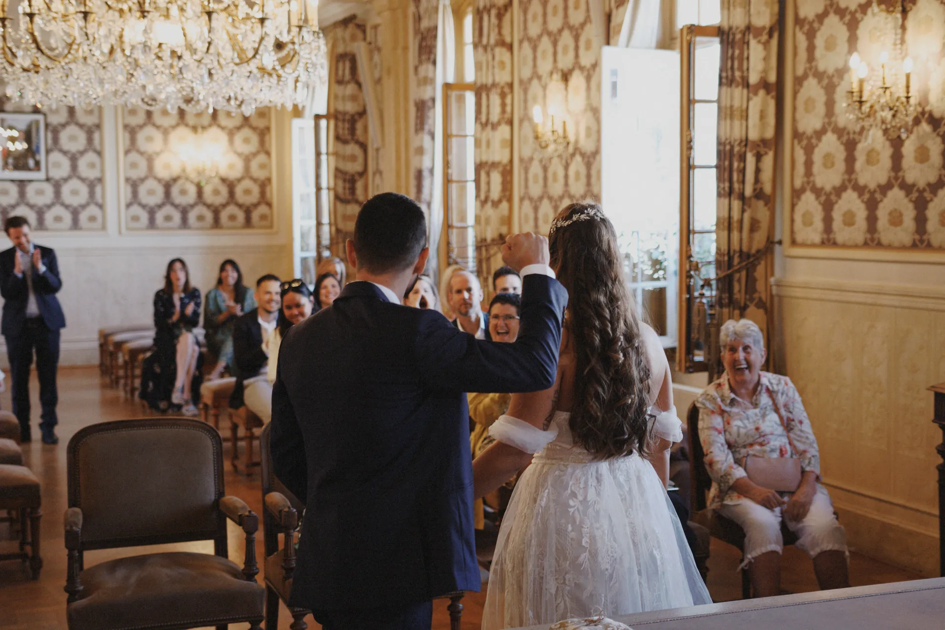 Les mariés se tiennent devant leurs invités assis dans une salle ornée de lustres et de murs à motifs. Capturé par Jeremy Dan, photographe et vidéaste de mariage basé à Sceaux, Hauts-de-Seine.