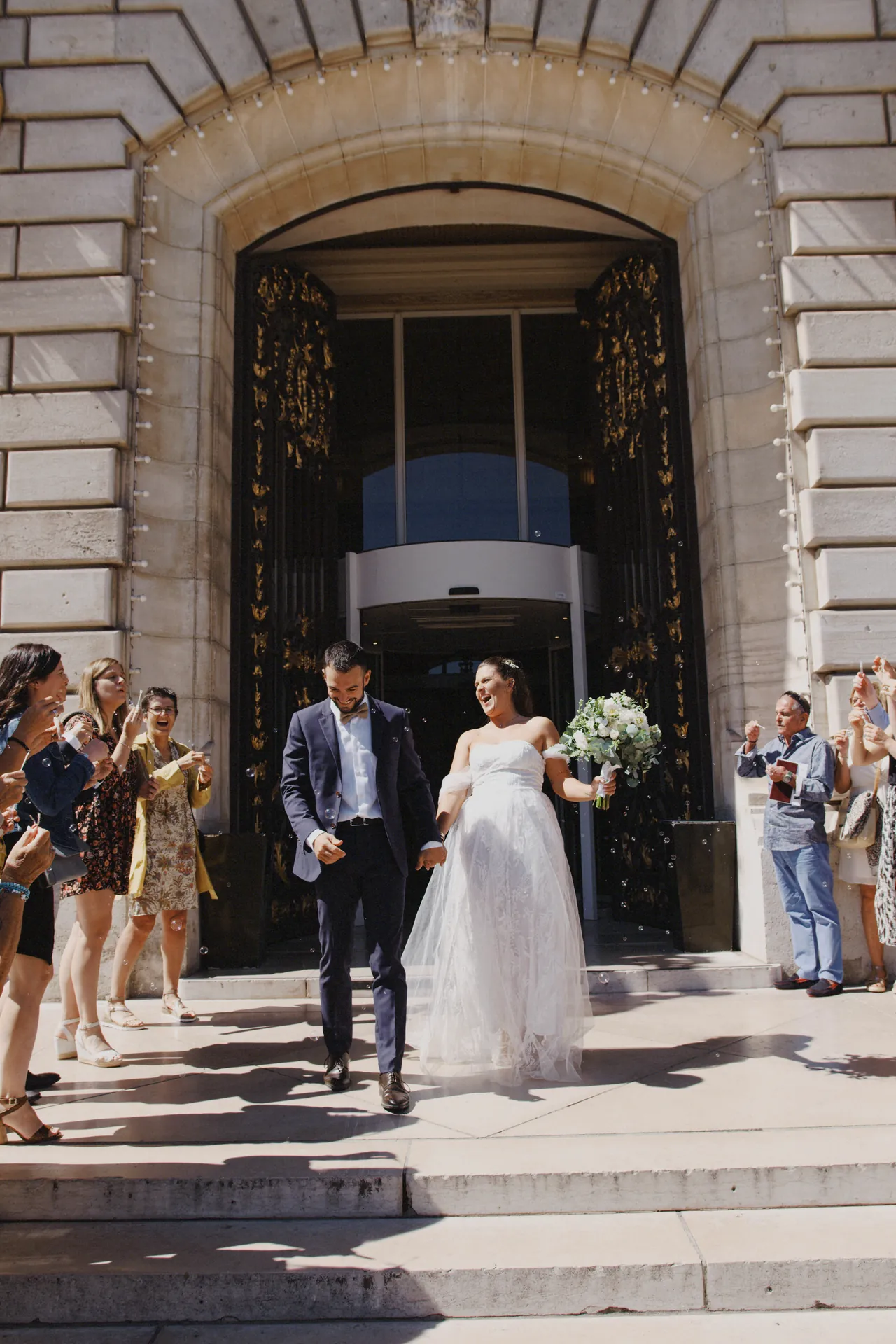 Des mariés descendent des marches à l’extérieur d’un grand bâtiment, entourés de gens qui applaudissent et prennent des photos. La mariée tient un bouquet, magnifiquement capturé par Jeremy Dan, photographe et vidéaste de mariage basé à Sceaux.