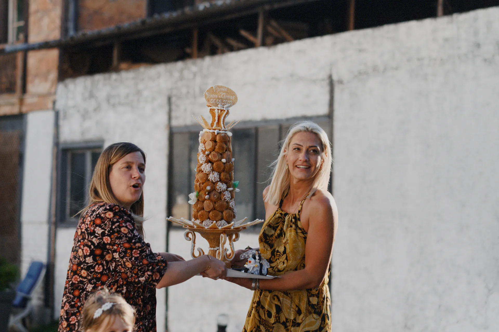 Deux femmes marchent et emmènent fièrement un grand croquembouche orné d'une garniture. Capturé par Jeremy Dan, photographe et vidéaste de mariage à Sceaux, Hauts-de-Seine.