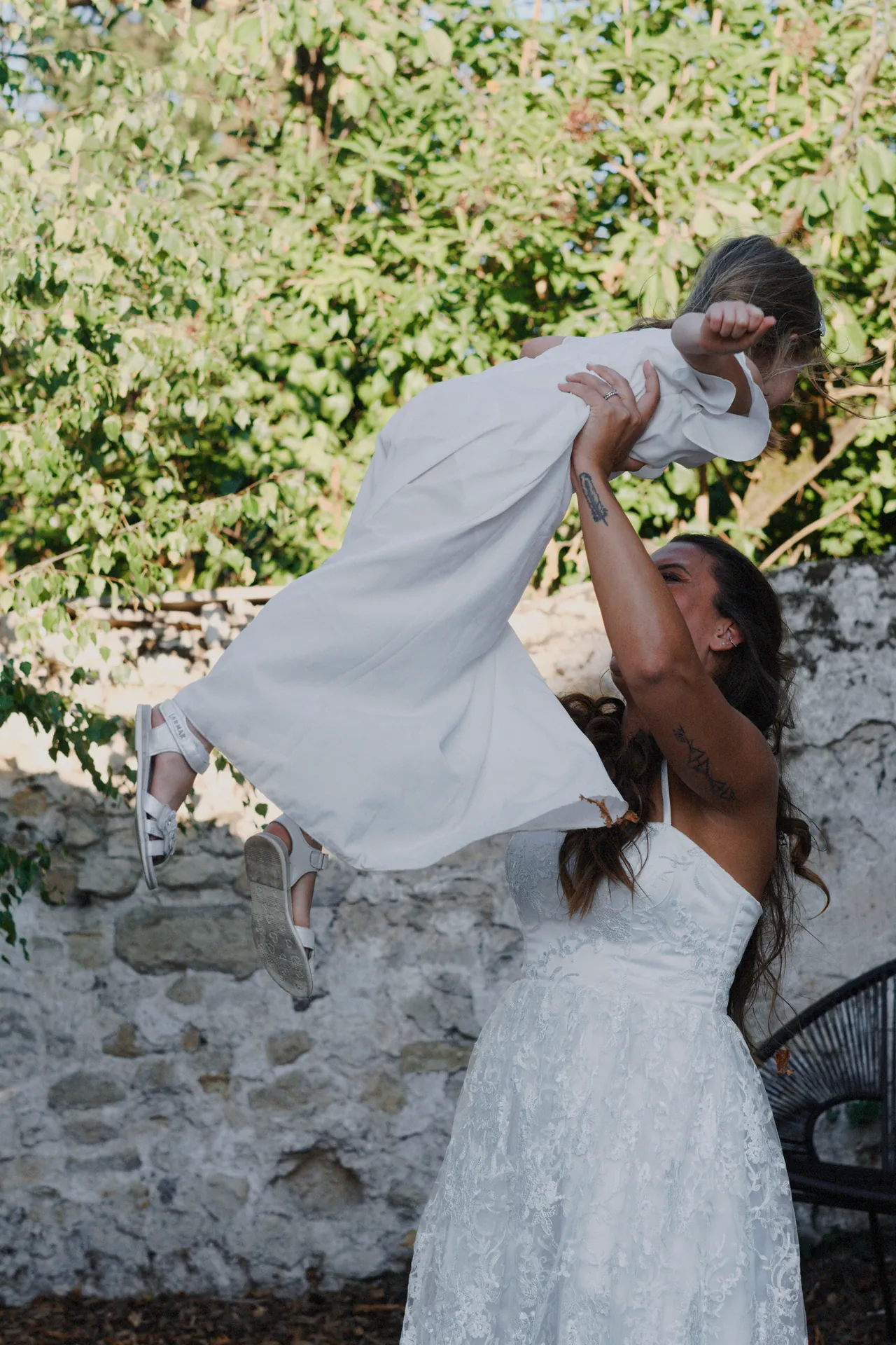 Une mariée en robe blanche porte joyeusement un enfant, également vêtu de blanc, sur fond de buissons verts et de mur de pierre. Capturé par Jeremy Dan, photographe et vidéaste de mariage à Sceaux, Hauts-de-Seine.
